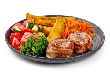Grilled bacon-wrapped pork medallions with potato wedges, carrot salad, and fresh vegetables on a black plate, isolated on white background.