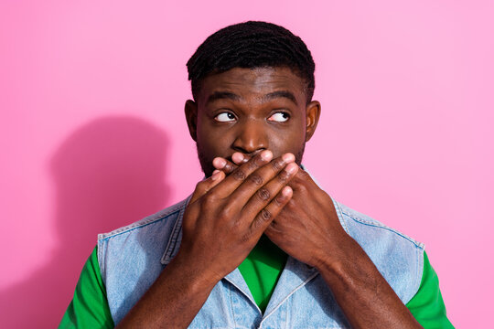 Young man with a playful expression covering his mouth against a pink background, wearing denim vest and green shirt