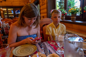 A woman in casual attire is absorbed in her smartphone while a boy in a yellow shirt watches attentively in a warm restaurant with rustic decor