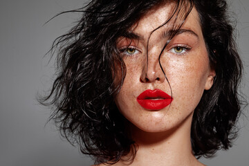 Portrait of a young woman with bold red lipstick, tousled black hair, and freckles, looking straight at the camera against a dark gray background