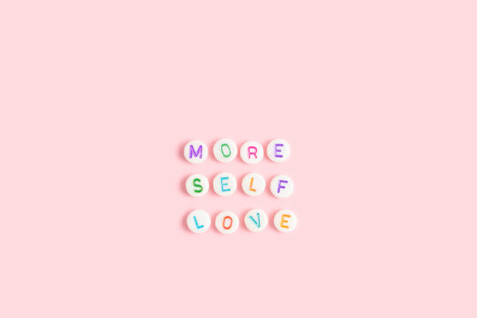 More self love. Quote made of white round beads with multicolored letters on a pink background.