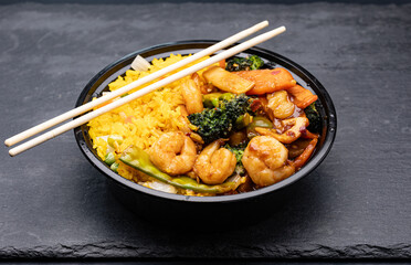 A Chinese food delivery dish featuring shrimp and Asian vegetables in garlic sauce, served with yellow fried rice in a disposable plastic bowl, accompanied by a pair of wooden chopsticks.