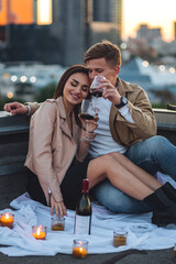 Romantic beautiful surprise on Saint Valentine's Day for a girlfriend. Loving young couple having dinner on a rooftop on sunset evening with candles, wine. Skyscrapers on background, urban cityscape