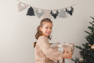 A little happy girl is sitting with present next to the Christmas tree. Christmas and New Year atmosphere with lights, coziness. The atmosphere of the holiday