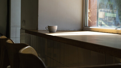 A coffee cup sits on a table near the window in a cozy café with wooden chairs. Hard sunlight.
