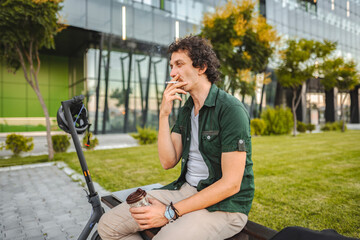 Man with curly hair sits on a park bench, smokes a cigar and rests