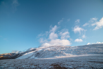 Lush sunlit low cloud on snow-capped mountain top at sunrise. Snowy stone hill in sunlight at early morning. High mountain in freshly fallen snow. Snow-covered stony peak and fluffy cloud in blue sky.