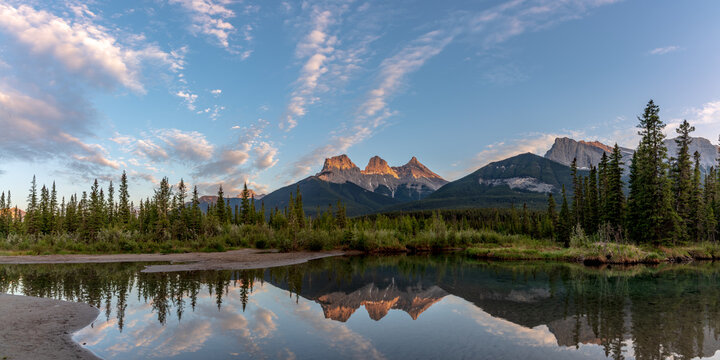 Three Sisters in Canmore seen at golden hour, sunset on blue sky day, afternoon with calm, peaceful reflection in water below famous, tourist, tourism mountains, area summer.	