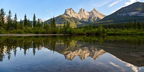 Three Sisters in Canmore seen at golden hour, sunset on blue sky day, afternoon with calm, peaceful reflection in water below famous, tourist, tourism mountains, area summer.	