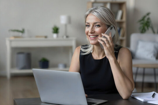 Cheerful happy middle aged entrepreneur woman talking on cellphone at table with laptop, using wireless online connection technology for work from home, smiling, enjoying mobile phone conversation
