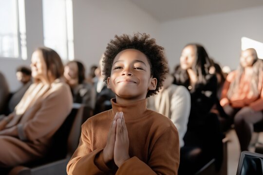 Hopeful african american boy with closed eyes and clasped hands praying in church during sunday service with congregation in background - Powered by Adobe