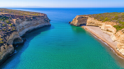 Fototapeta premium A serene coastal view featuring a turquoise bay surrounded by rocky cliffs and a sandy beach under a clear blue sky.