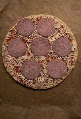 A pizza with salami and cheese on a brown paper. The pizza is ready to be baked