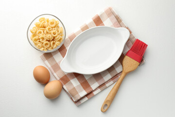 Ceramic casserole, raw pasta, eggs and brush on white table, flat lay