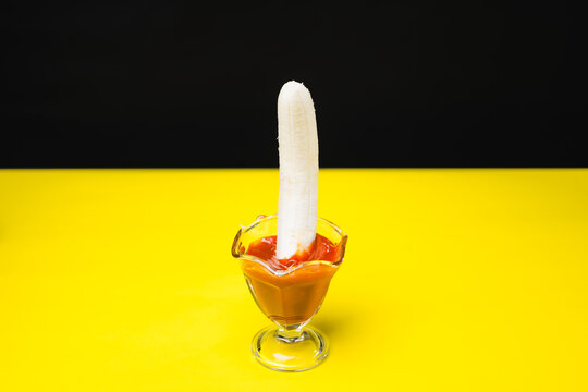 A banana piece dipped into a glass container of ketchup on a yellow and black background