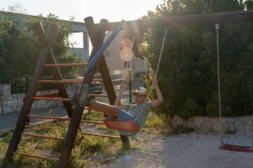 Young boy swinging on red swing set in sunny outdoor playground, wearing cap and casual outfit. Concept of carefree childhood, outdoor fun, and active lifestyle on warm summer day. High quality photo