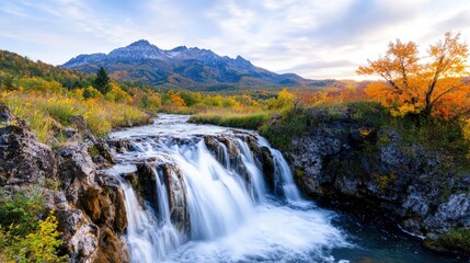 Obraz premium Serene autumn waterfall cascading down rocky cliffs into a pool, with vibrant fall foliage and snow-capped mountains in the background.