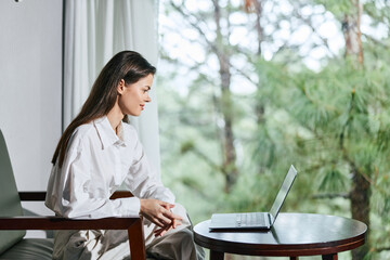 Young woman in casual white shirt sitting by a laptop, engaged in remote work, showcasing focus and...