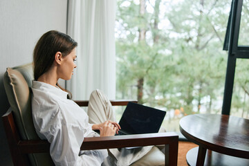 Young professional woman working on a laptop in a cozy living room with natural light, reflecting focus and tranquility amidst a green outdoor background
