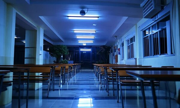 A dimly lit corridor in a school with empty tables and chairs, creating a quiet atmosphere.