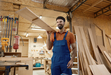 Smiling woodworker carrying timber plank on shoulder over workshop