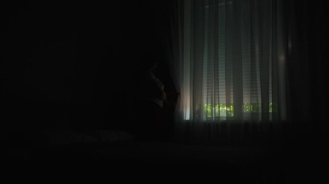 Woman raising electric window shutters with button in dark room in morning, wide shot. Letting daylight into bedroom during morning routine.