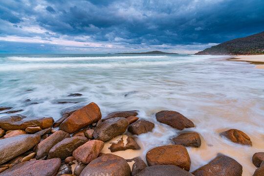 A dark overcast sky advancing over rough waves rolling over rocks on a beach