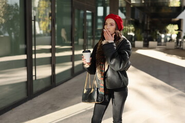 A stylish woman in a red beanie and leather jacket sips coffee while strolling through the vibrant city