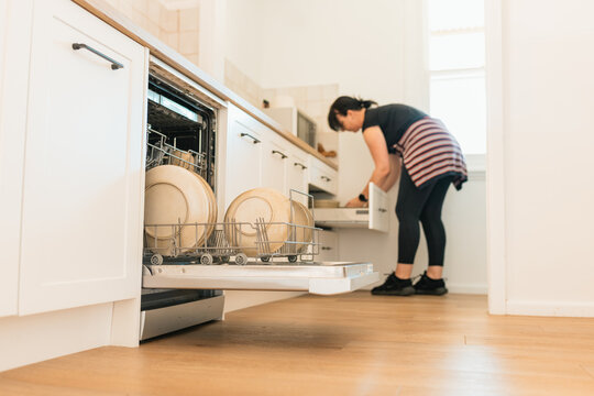 Woman unloading dishwasher in bright kitchen with clean plates and cookware on wooden floor
