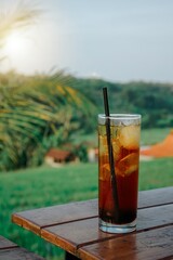 Full fresh iced tea in a glass, with a straw and ice cubes in it, a glass of iced tea in a glass against a nature background, iced tea on a wooden table. 