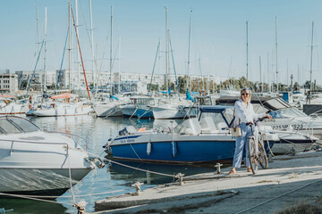 Fototapeta premium Woman enjoys bike ride along waterfront, Marina surroundings. She is wearing a white shirt and blue jeans, and she has a handbag with her. Capturing outdoor bike ride by waterfront.
