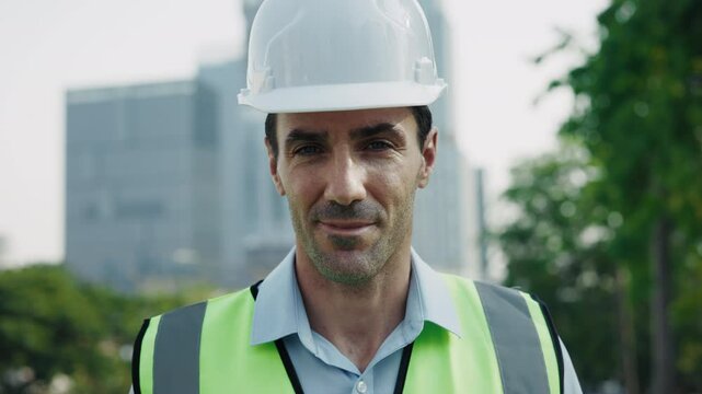 Portrait of confident engineer looking at camera standing at construction site. Smiling man in hardhat and vest with positive facial expression reflects satisfaction with work