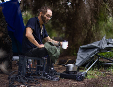 homeless man sitting on milk crate near butane stove in front of tarpaulin camp