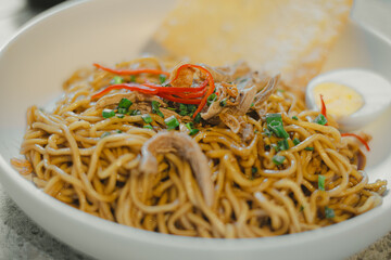 Indonesian Fried Noodles with toppings: Boiled egg, crackers, chili, and leeks in a white bowl on stone table