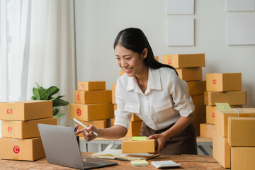 Smiling Entrepreneur Managing Online Orders: A cheerful young Asian woman efficiently manages her thriving e-commerce business, surrounded by stacks of shipping boxes and using a laptop.