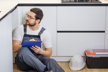 serious adult craftsman writing in clipboard while sitting on floor in kitchen