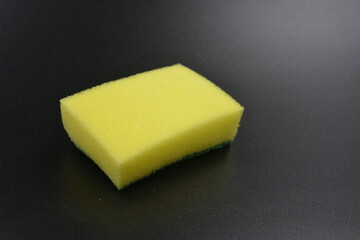 A small yellow sponge with a green rough dishwashing surface placed on a black, matte plastic background.