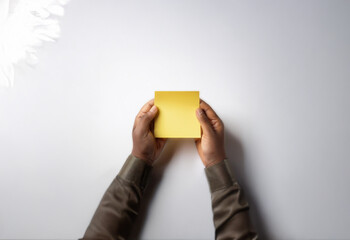 A pair of hands hold a blank yellow sticky note against a white background.