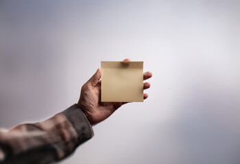 A hand holds a blank yellow sticky note against a blurred background.