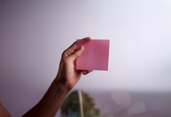 A hand holds a pink sticky note against a white background, with a blurred green plant in the background.
