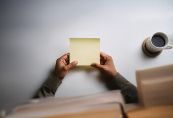 A person's hands hold a blank yellow sticky note against a white background. A cup of coffee sits on a coaster to the right. Books lay on the surface below the person's hands.