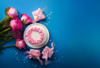 A single-tiered white cake with pink frosting sits on a white plate surrounded by pink roses, pink presents, and pink sprinkles on a blue background.