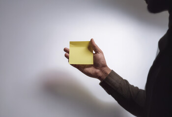 A hand holds a blank yellow sticky note against a white background.