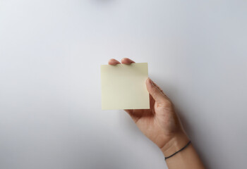 A hand holds a blank yellow sticky note against a plain white background.