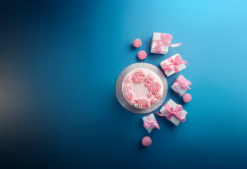 Fototapeta premium A pink frosted cake with decorative piping sits on a white cake board surrounded by wrapped gifts and pink candles on a blue background.