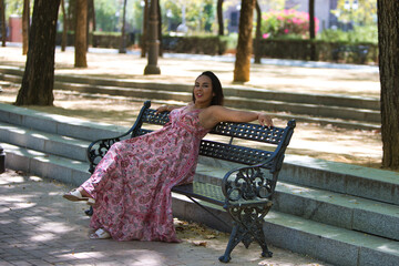 a young, beautiful brunette woman is sitting on a park bench, her arms resting on the backrest, her...