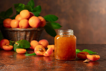 Apricot jam in glass jar and fresh fruits on a brown table.