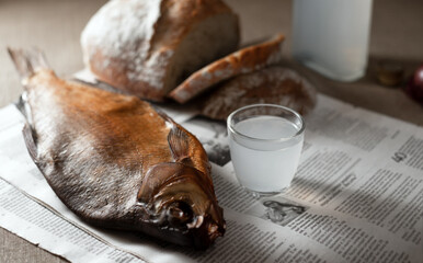 Still life of traditional Eastern European food featuring a glass and carafe of vodka, smoked fish, dark rye bread, and sliced purple onion arranged on a rustic newspaper background	