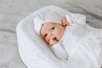 A baby is laying on a white blanket with a white hat on. The baby is looking at the camera and he...