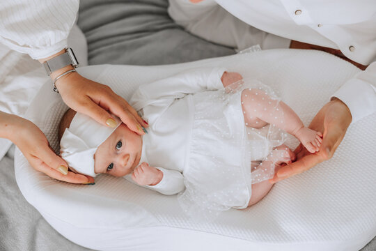 A baby is laying on a white blanket with a white dress on. A woman is holding the baby's head